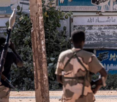 Trois hommes en tenue militaire et armés se trouvent dans une rue déserte. L’un porte un lance-roquettes sur l’épaule, un autre a une arme en bandoulière, et le troisième est assis au sol avec une mitrailleuse.