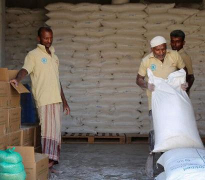 Trois hommes travaillant pour le Programme alimentaire mondial (PAM), dont l’un soulève un sac de riz. En arrière-plan, plusieurs sacs de riz empilés.