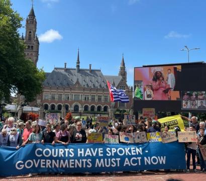 Manifestation devant le Palais de la Paix à La Haye, aux Pays-Bas. Une grande bannière bleue portée par des manifestants affiche le message : “Courts have spoken – Governments must act now”. Les participants tiennent également d’autres pancartes sur le climat et la justice environnementale. Un écran géant diffuse le discours d'une intervenante, et des caméras de médias sont visibles au premier plan. L’événement se déroule sous un ciel bleu, dans une ambiance pacifique mais engagée.