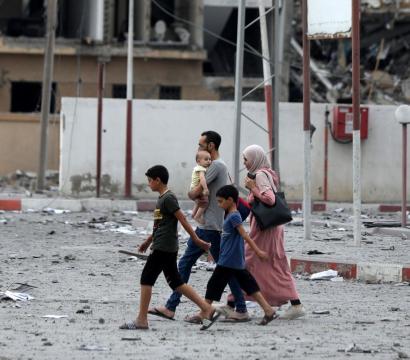 Une famille composée de deux adultes et quatre enfants traverse une rue jonchée de débris et de papiers, dans une zone fortement endommagée par des destructions. En arrière-plan, des bâtiments partiellement effondrés témoignent de récentes violences. Le père porte un bébé dans les bras, tandis que les autres enfants marchent à côté de leur mère, qui est voilée. L’atmosphère est tendue, marquée par les traces visibles d’un conflit.