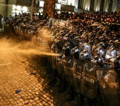 Un groupe compact de policiers antiémeute, protégés par des boucliers transparents et casques, fait face à des manifestants de nuit. Un policier utilise un gaz irritant ou spray poivre contre un manifestant replié en avant. La scène se déroule sur une rue pavée, dans une ambiance tendue et violente, avec une forte présence policière en arrière-plan.