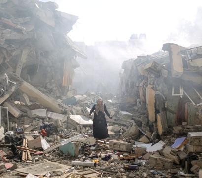 Une femme marche seule au milieu des décombres d’immeubles effondrés, entourée de gravats, de poutres brisées et de fragments de béton. Une fumée épaisse s’élève à l’arrière-plan, suggérant une récente explosion ou attaque. L’atmosphère est lourde, marquée par la destruction et le chaos.