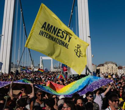 Une foule nombreuse participe à une marche ou manifestation sur un pont, portant des drapeaux arc-en-ciel et un grand drapeau jaune d’Amnesty International. Les participants sourient, brandissent des pancartes et des bannières multicolores sous un ciel bleu clair. L’événement semble festif et engagé, promouvant les droits humains et la cause LGBTQIA+.
