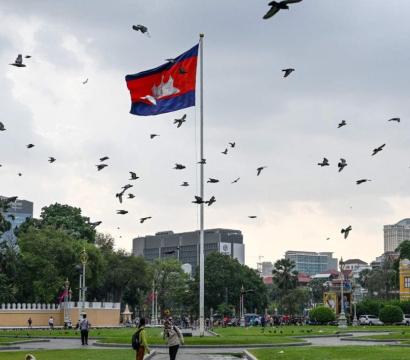 « Place publique à Phnom Penh, Cambodge, avec un grand mât portant le drapeau cambodgien flottant au vent. De nombreux oiseaux volent dans le ciel nuageux. Des passants se promènent sur les pelouses bien entretenues, entourées de bâtiments coloniaux colorés, d’arbres et de lampadaires dorés. En arrière-plan, on aperçoit des immeubles modernes.
