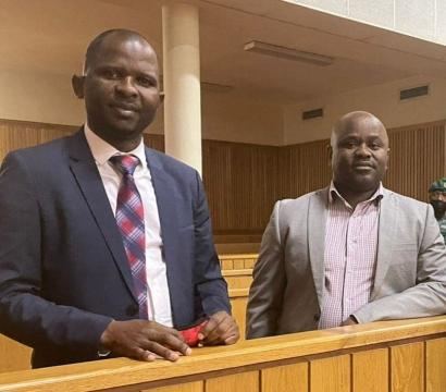 « Deux hommes en costume se tiennent debout dans un tribunal, derrière une barrière en bois. L’un porte une cravate à motifs rouges et bleus, tandis que l’autre est en chemise à carreaux et veste grise. En arrière-plan, deux gardes en uniforme vert surveillent la salle d’audience.
