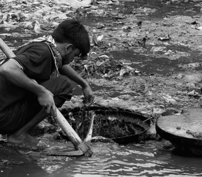 Un homme accroupi travaille dans des eaux sales et boueuses, manipulant un outil semblable à une pioche près d’une bouche d’égout ouverte. L’environnement est jonché de détritus et reflète des conditions de travail dangereuses et insalubres.