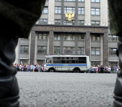 Vue au ras du sol entre deux bottes de militaire ou de policier, face au bâtiment de la Douma d’État en Russie. Une foule de personnes se tient en file le long du trottoir, à côté d’un bus de police stationné. La scène suggère une manifestation ou un rassemblement surveillé devant une institution gouvernementale.