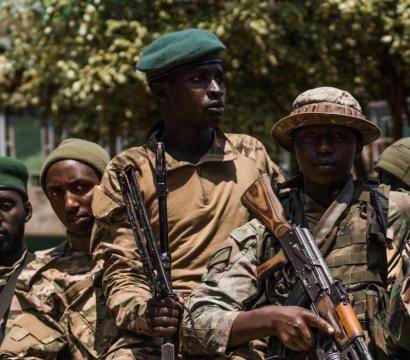 Groupe de soldats armés portant des uniformes militaires et des bérets verts ou noirs. Certains tiennent des fusils d’assaut, tandis qu’ils se tiennent serrés les uns contre les autres, semblant prêts à une opération. En arrière-plan, on aperçoit des arbres et un bâtiment.
