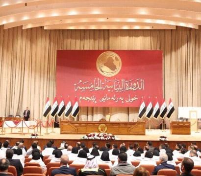 « Vue d’ensemble d’une séance parlementaire dans un grand hémicycle en Irak. Des parlementaires sont assis face à une estrade ornée de multiples drapeaux irakiens et d’un grand panneau rouge portant des inscriptions en arabe et un emblème doré représentant la carte du pays. Plusieurs responsables se tiennent debout sur la scène, tandis que des caméras et agents de sécurité sont présents dans la salle. 