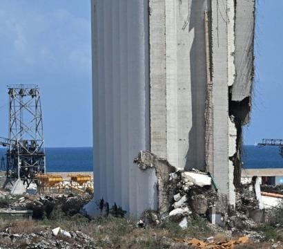 Vue sur un silo gravement endommagé à proximité du port, avec des gravats et des structures métalliques en arrière-plan. À l'horizon, on aperçoit la mer Méditerranée sous un ciel bleu. Des drapeaux libanais sont visibles dans le coin inférieur droit. 