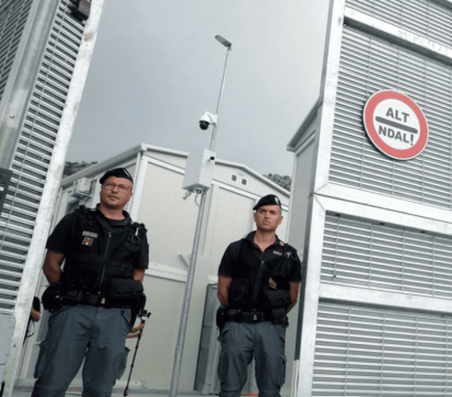 « Deux agents de sécurité en uniforme noir se tiennent debout devant une haute clôture métallique munie de caméras de surveillance. Un panneau circulaire rouge et blanc portant l'inscription "ALT NDAL!" est visible sur la grille. L'arrière-plan montre des structures modulaires blanches sous un ciel gris. 