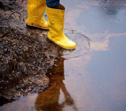 Une personne portant des bottes jaunes en caoutchouc se tient sur un sol boueux et souillé par une nappe de pétrole, dans une zone polluée par des hydrocarbures.