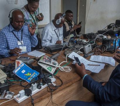 Un groupe de journalistes et techniciens assis autour d’une grande table en bois, équipée de téléphones fixes, câbles, ordinateurs portables, enregistreurs et autres matériels audio. Tous portent des casques sur les oreilles et consultent des téléphones ou des documents, concentrés sur leur travail. L’ambiance reflète l’intensité d’une salle de presse en pleine activité.
