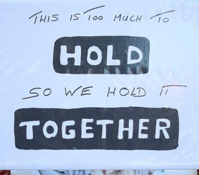 Une pancarte tenue par une personne lors d’une manifestation. Le panneau blanc, bordé de rouge, porte un message manuscrit en anglais : « This is too much to HOLD so we hold it TOGETHER ». Les mots « HOLD » et « TOGETHER » sont mis en évidence en lettres majuscules blanches sur fond noir. En arrière-plan flou, d’autres manifestants et drapeaux palestiniens apparaissent.