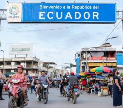 Plusieurs personnes circulent à scooter devant un panneau "Bienvenue en Équateur"